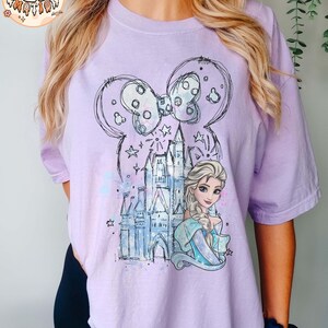 Puede incluir: Una camiseta morada con un boceto de un castillo, un lazo y una princesa con cabello rubio. La princesa lleva un vestido azul y una corona. El texto "Orchid" es visible en la esquina superior derecha de la imagen.