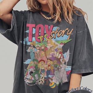 Peut inclure: Un t-shirt gris anthracite oversize avec une impression graphique portant le texte "TOY story" en rose et orange. Le motif pr&eacute;sente un collage de divers personnages f&eacute;minins dans des tenues color&eacute;es, un ballon jaune et bleu, et un appareil photo. Le coin inf&eacute;rieur droit inclut le texte "Comfort Colors Pepper".