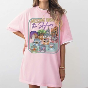 May include: Pink t-shirt with a graphic featuring the text "WELCOME ABOARD the Skyliner" and illustrations of Disney characters in gondolas. The design includes images of Epcot and other park landmarks.