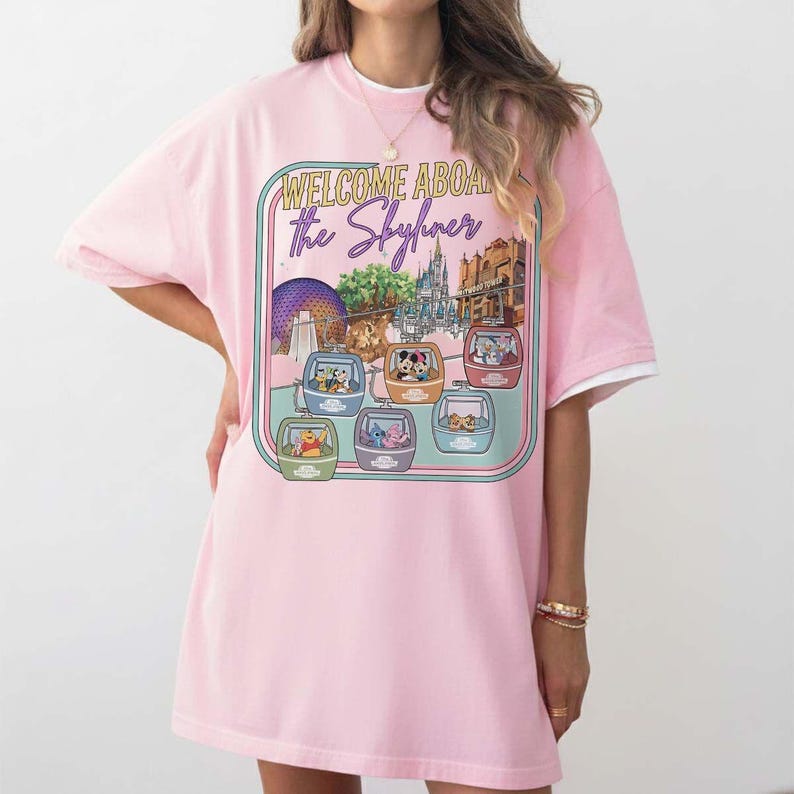 May include: Pink t-shirt with a graphic featuring the text "WELCOME ABOARD the Skyliner" and illustrations of Disney characters in gondolas. The design includes images of Epcot and other park landmarks.