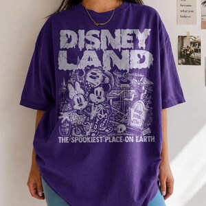 May include: A purple t-shirt with the text "DISNEY LAND" in a dripping font. The graphic features Mickey and Minnie Mouse, a graveyard scene, and the words "THE SPOOKIEST PLACE ON EARTH."