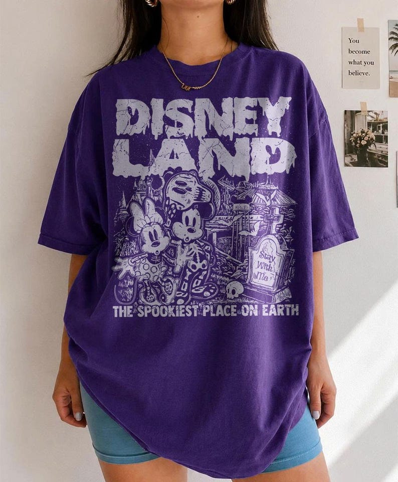 May include: A purple t-shirt with the text "DISNEY LAND" in a dripping font. The graphic features Mickey and Minnie Mouse, a graveyard scene, and the words "THE SPOOKIEST PLACE ON EARTH."
