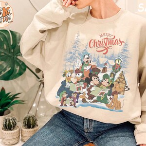 Może przedstawiać: Beżowa bluza z nadrukiem świątecznym przedstawiającym Myszki Miki, Minnie, Donalda, Goofy'ego i Pluto. Nadruk zawiera napis "Merry Christmas".