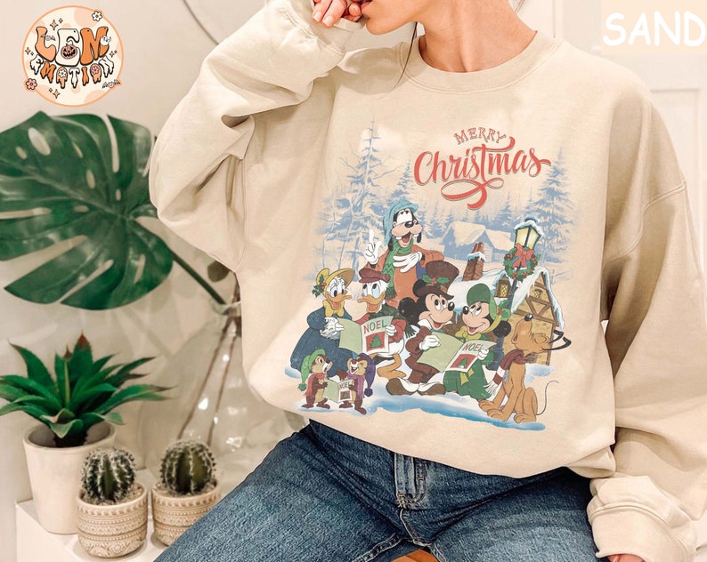 Może przedstawiać: Beżowa bluza z nadrukiem świątecznym przedstawiającym Myszki Miki, Minnie, Donalda, Goofy'ego i Pluto. Nadruk zawiera napis "Merry Christmas".