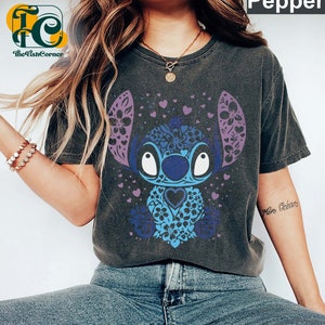 May include: A dark gray t-shirt with a blue and purple Stitch character design. The character is surrounded by hearts and flowers. The text "More Dreams" is visible on the right sleeve.