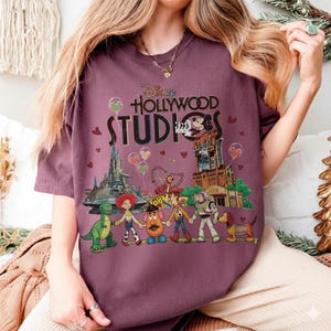 Op de afbeelding: Een bordeauxrood T-shirt met de tekst "Disney Hollywood Studios" en een afbeelding van Disney-personages en parkattracties. Het ontwerp bevat Mickey Mouse, Toy Story-personages en de Tower of Terror.