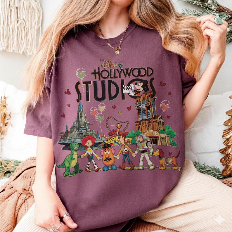 Op de afbeelding: Een bordeauxrood T-shirt met de tekst "Disney Hollywood Studios" en een afbeelding van Disney-personages en parkattracties. Het ontwerp bevat Mickey Mouse, Toy Story-personages en de Tower of Terror.