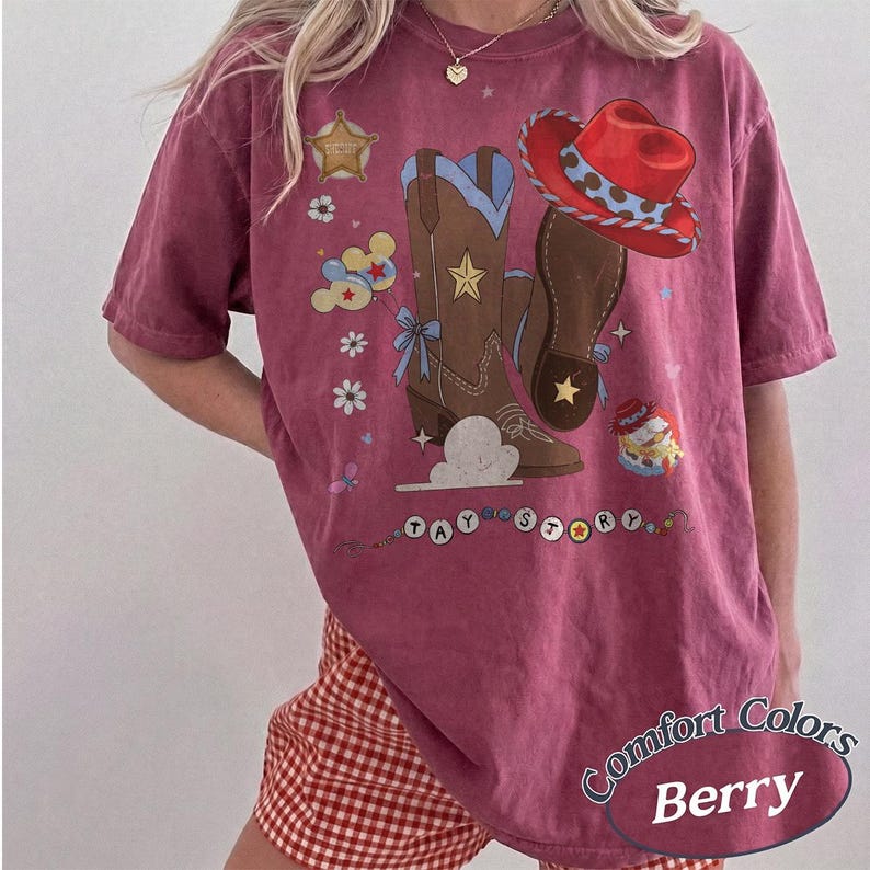 Pu&ograve; includere: Una t-shirt color bacca con una stampa grafica di stivali da cowboy marroni, un cappello da cowboy rosso, una stella da sceriffo dorata e palloncini colorati. Il testo "TAY STORY" &egrave; stampato sotto gli stivali. Girocollo e maniche corte.