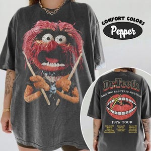Könnte beinhalten: Ein anthrazitfarbenes T-Shirt mit einer Grafik der Muppet-Figur Animal, die Trommelstöcke hält. Die Rückseite zeigt das Logo von Dr. Teeth and the Electric Mayhem mit einem Mund-Motiv, dem Text 1976 TOUR und einer Liste von Städten.