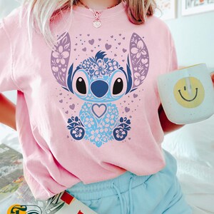 May include: A pink t-shirt with a blue and purple floral Stitch design. The design features a blue Stitch character with a heart in its chest. The shirt is worn by a person with blue shorts and a book in their hand.