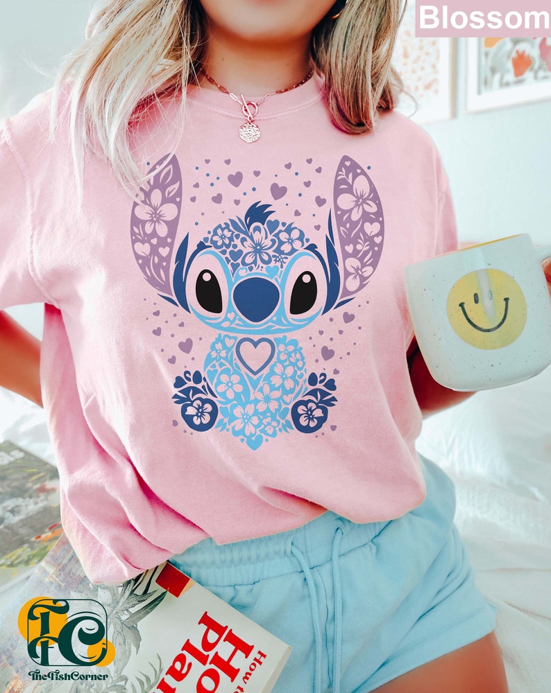 May include: A pink t-shirt with a blue and purple floral Stitch design. The design features a blue Stitch character with a heart in its chest. The shirt is worn by a person with blue shorts and a book in their hand.