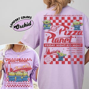 K&ouml;nnte beinhalten: Ein helllila T-Shirt mit dem Pizza Planet-Logo und Alien-Charakteren. Auf der R&uuml;ckseite des Shirts befindet sich ein rot-wei&szlig; kariertes Muster mit dem Text "Friday Night Kid Night". Das Shirt ist mit "Comfort Colors Orchid" gekennzeichnet.