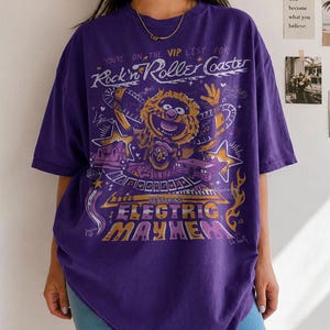 Puede incluir: Camiseta morada con un dise&ntilde;o gr&aacute;fico que incluye el texto "Rock'n Roller Coaster" y "Electric Mayhem", junto con un personaje de dibujos animados y estrellas.