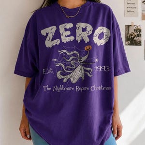 May include: A purple short-sleeved t-shirt featuring a graphic of the character Zero from The Nightmare Before Christmas. The design includes the text ZERO, Est. 1993, and The Nightmare Before Christmas, with a spiderweb and pumpkin motif.