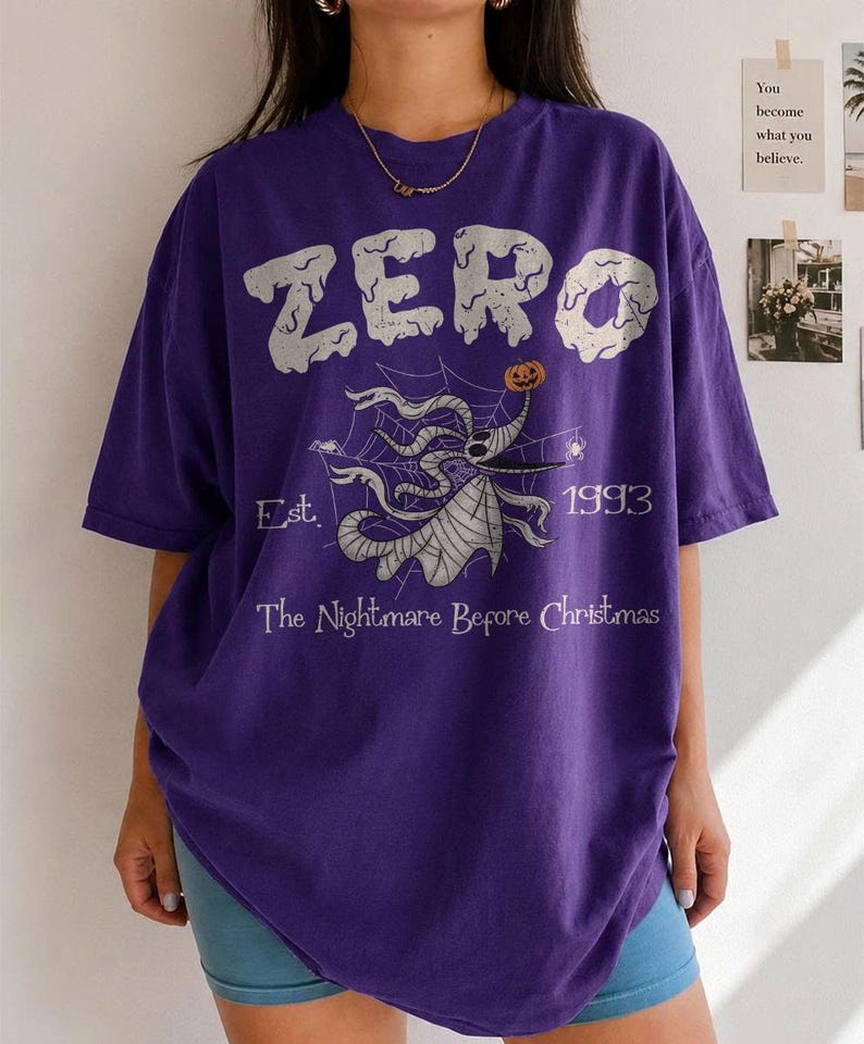 May include: A purple short-sleeved t-shirt featuring a graphic of the character Zero from The Nightmare Before Christmas. The design includes the text ZERO, Est. 1993, and The Nightmare Before Christmas, with a spiderweb and pumpkin motif.