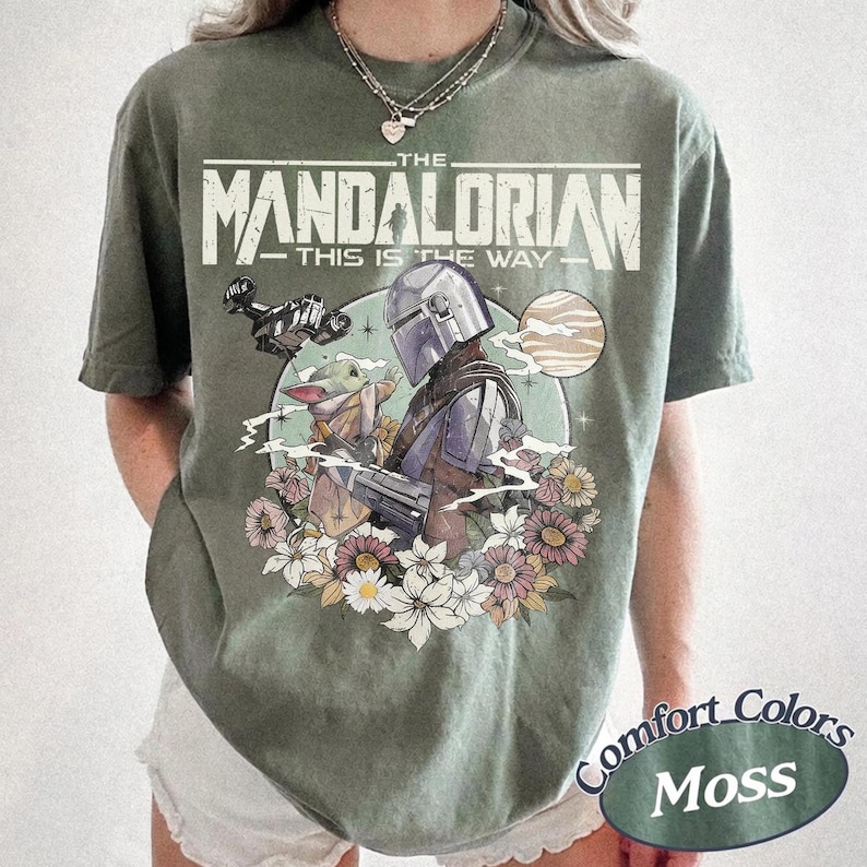 May include: A moss green short-sleeve t-shirt featuring a graphic print of The Mandalorian and Grogu. The text reads THE MANDALORIAN THIS IS THE WAY. The design includes a spaceship, a planet, and a cluster of colorful flowers at the bottom.