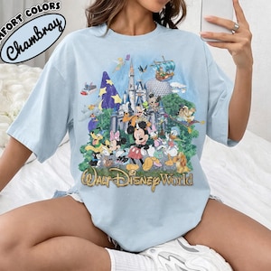 May include: A light blue t-shirt featuring a graphic print of the Walt Disney World castle surrounded by various cartoon characters including Mickey Mouse, Minnie Mouse, Donald Duck, and Goofy, with the text Walt Disney World at the bottom.