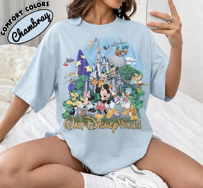 May include: A light blue t-shirt featuring a graphic print of the Walt Disney World castle surrounded by various cartoon characters including Mickey Mouse, Minnie Mouse, Donald Duck, and Goofy, with the text Walt Disney World at the bottom.