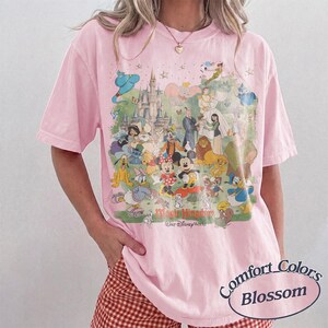 May include: A light pink short-sleeve t-shirt featuring a large, colorful graphic print of various animated characters and a castle. The text Magic Kingdom and Walt Disney World is printed below the characters. The shirt has a crew neckline.