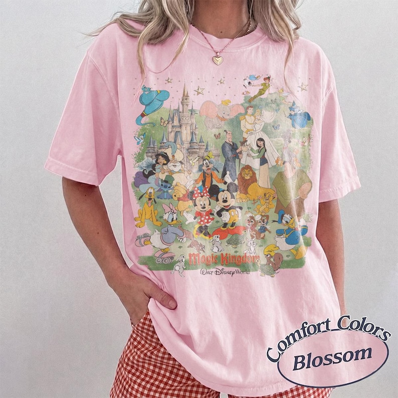 May include: A light pink short-sleeve t-shirt featuring a large, colorful graphic print of various animated characters and a castle. The text Magic Kingdom and Walt Disney World is printed below the characters. The shirt has a crew neckline.