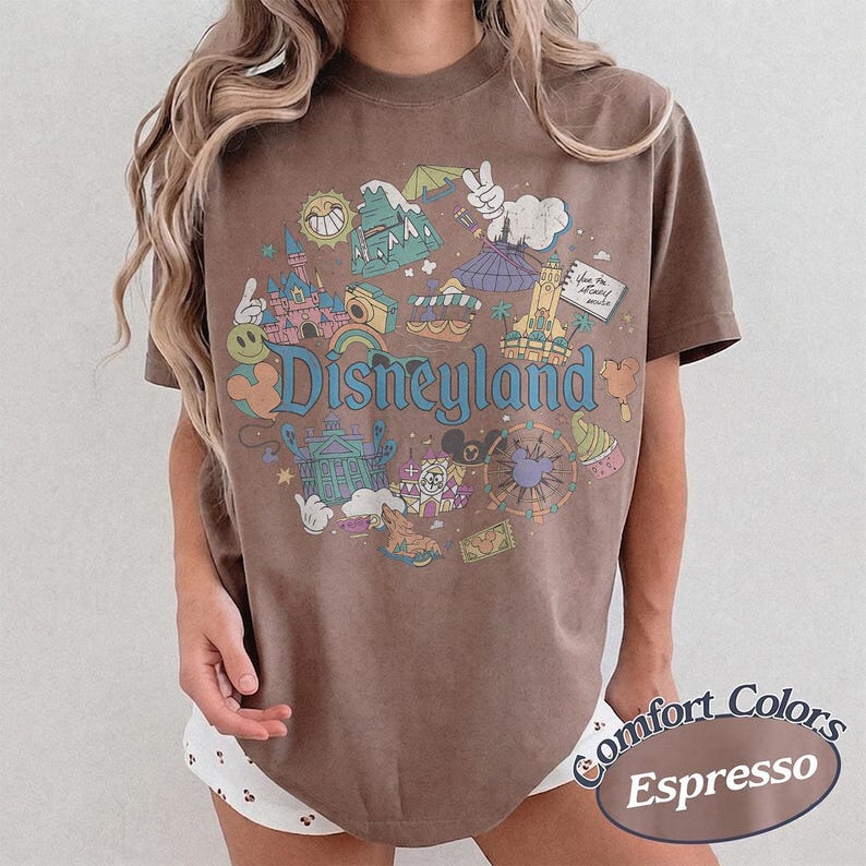 May include: Brown t-shirt with a Disneyland graphic. The graphic features various cartoon elements, including a castle, a sun, and Mickey Mouse. The text "Disneyland" is in a colorful, playful font. The shirt is labeled "Comfort Colors Espresso".
