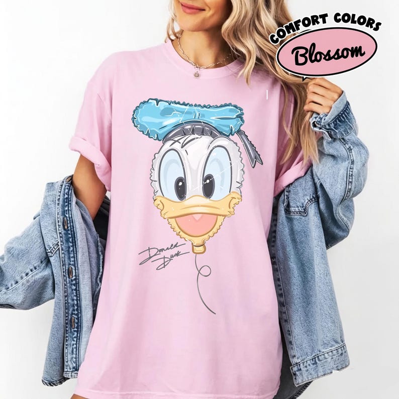 Pode incluir: Uma t-shirt de manga curta rosa claro com um gr&aacute;fico do rosto do Pato Donald desenhado como um bal&atilde;o com um la&ccedil;o azul no topo. O texto Donald Duck est&aacute; escrito em cursiva preta abaixo do gr&aacute;fico. A t-shirt tem uma gola redonda.