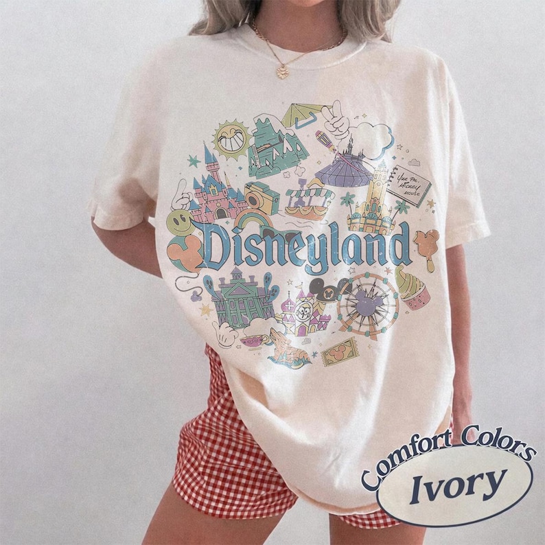 May include: Ivory-colored t-shirt with a Disneyland graphic. The graphic features various park icons, including a castle, a Ferris wheel, and the Disneyland logo in blue. The shirt is labeled "Comfort Colors" and "Ivory".