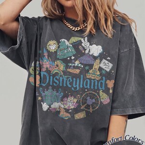 May include: Dark gray t-shirt with a colorful Disneyland graphic. The design includes various park icons, the Disneyland logo, and the words "Comfort Colors Pepper". The shirt has short sleeves and a relaxed fit.