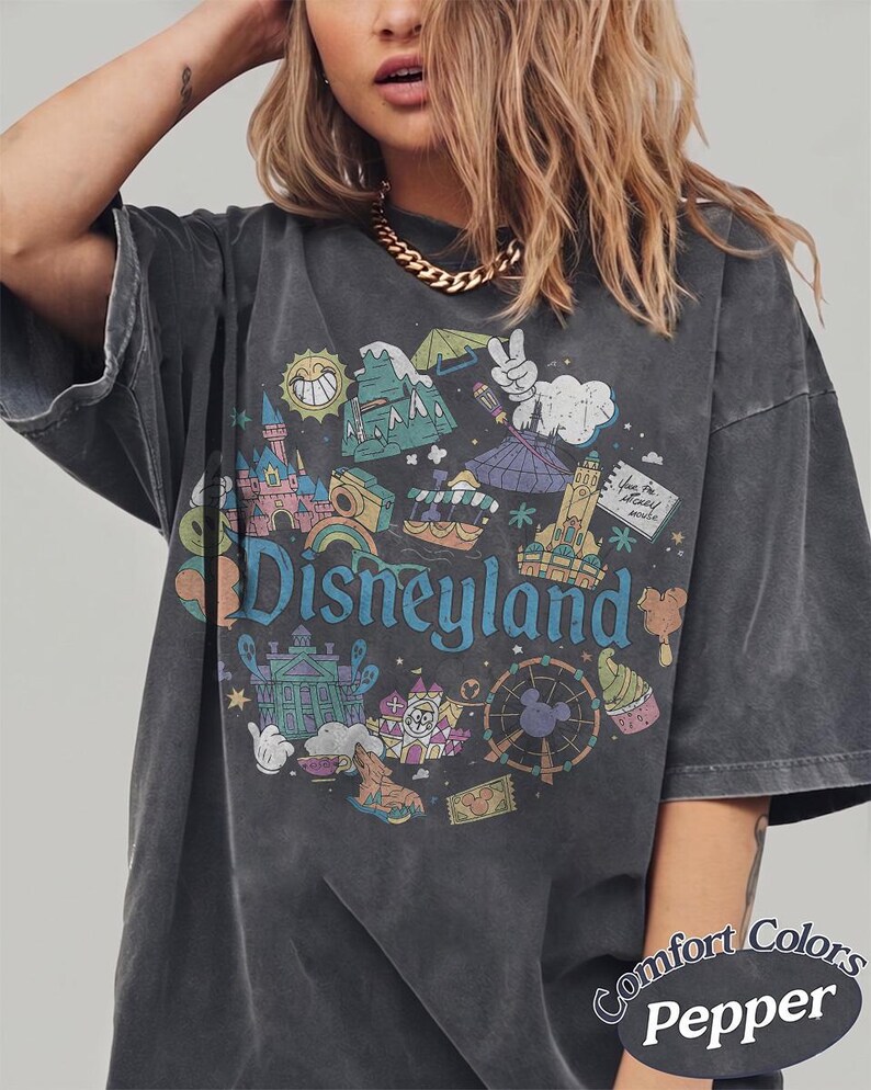 May include: Dark gray t-shirt with a colorful Disneyland graphic. The design includes various park icons, the Disneyland logo, and the words "Comfort Colors Pepper". The shirt has short sleeves and a relaxed fit.