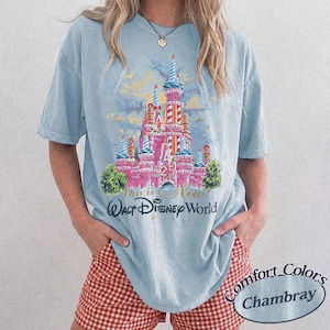 May include: Light blue t-shirt featuring a pink castle graphic with the text "This is Now! Walt Disney World 1997". The shirt has short sleeves and a crew neck. The model is wearing red and white checkered shorts.