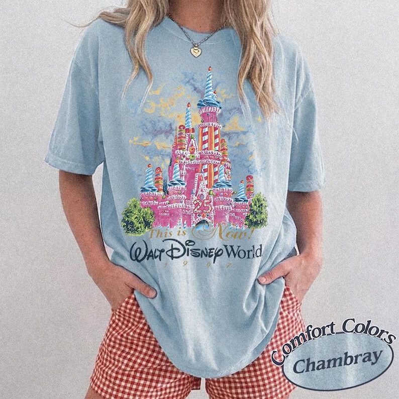 May include: Light blue t-shirt featuring a pink castle graphic with the text "This is Now! Walt Disney World 1997". The shirt has short sleeves and a crew neck. The model is wearing red and white checkered shorts.