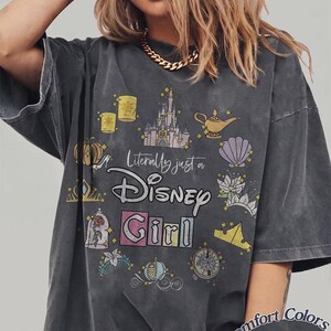 Op de afbeelding: Donkergrijs T-shirt met de tekst "Literally just a Disney Girl" en diverse Disney-thema afbeeldingen. Het shirt heeft een distressed, vintage look en de woorden "Comfort Colors Pepper" staan rechtsonder.