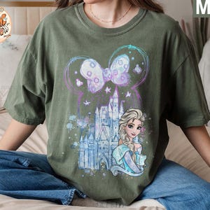 Puede incluir: Una camiseta verde con un dise&ntilde;o de acuarela que presenta un castillo, un lazo y una princesa con cabello rubio. La princesa lleva un vestido azul y una corona en la cabeza.