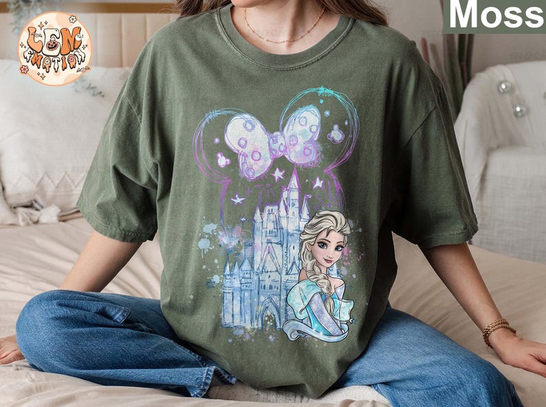 Puede incluir: Una camiseta verde con un dise&ntilde;o de acuarela que presenta un castillo, un lazo y una princesa con cabello rubio. La princesa lleva un vestido azul y una corona en la cabeza.