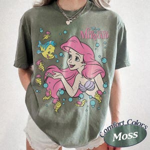 May include: A moss green short-sleeve t-shirt featuring a graphic of Ariel from The Little Mermaid with pink hair, a purple seashell top, and Flounder. The design includes small yellow seahorses and blue bubbles, with the text The Little Mermaid.