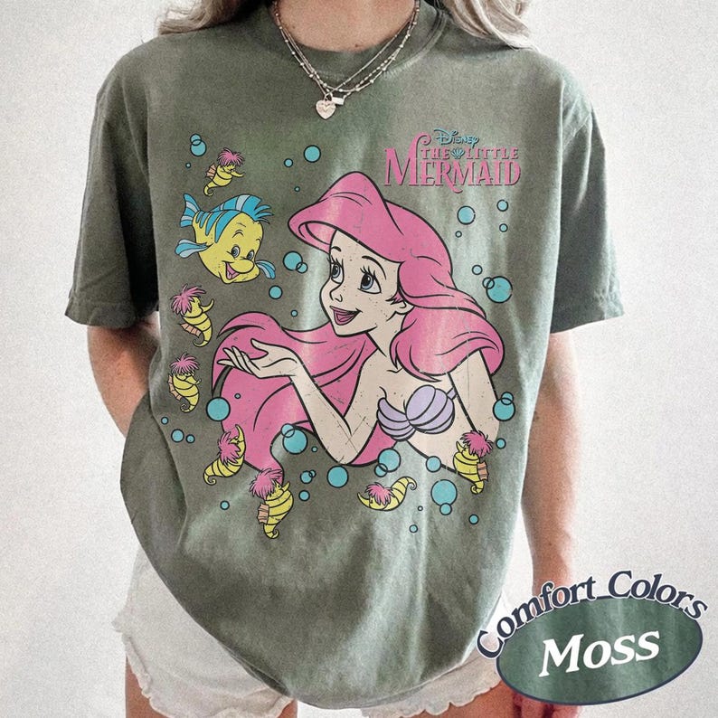 May include: A moss green short-sleeve t-shirt featuring a graphic of Ariel from The Little Mermaid with pink hair, a purple seashell top, and Flounder. The design includes small yellow seahorses and blue bubbles, with the text The Little Mermaid.