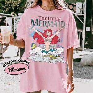 May include: A light pink t-shirt featuring a graphic print of Ariel from The Little Mermaid with red hair, a seashell top, and a green tail. The design includes Flounder, Sebastian, and the text The Little Mermaid and Disney in a vintage style.