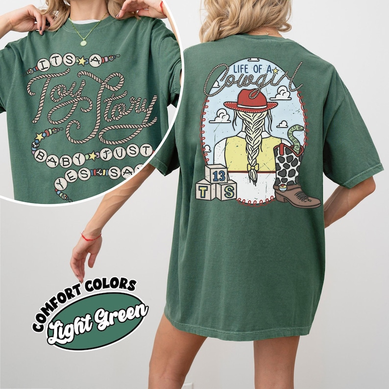 May include: A light green t-shirt featuring a graphic on the back of a person with a braided ponytail wearing a red cowboy hat, a yellow shirt, and a cow-print boot with a snake, plus blocks labeled 13 TS and the text Life of a Cowgirl.