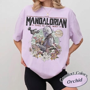 May include: A light purple t-shirt featuring a graphic print of The Mandalorian and Grogu. The text reads THE MANDALORIAN THIS IS THE WAY. The design includes a spaceship, a planet, and various flowers surrounding the characters in a circular frame.