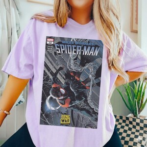 Op de afbeelding: Een lichtpaars T-shirt met een stripboekcover van Spider-Man. De cover toont Spider-Man in een zwart en rood pak, met de tekst "Miles Morales: Spider-Man" en "Gang War is Coming."