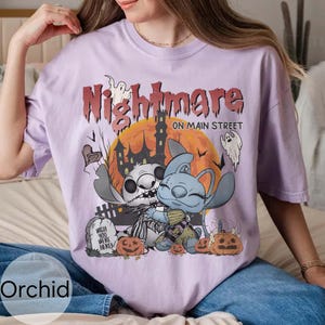 Puede incluir: Una camiseta lila con un gr&aacute;fico de Halloween. El dise&ntilde;o incluye el texto "Nightmare on Main Street" sobre una imagen de dibujos animados de dos personajes, una casa embrujada, calabazas y fantasmas. La camiseta tiene un corte holgado.
