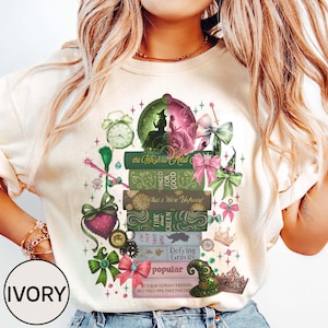以下が含まれることがあります： ファンタジーをテーマにしたデザインのアイボリー色のTシャツ。「the Wizard and」や「Defying Gravity」などのテキストが書かれた本が積み重ねられ、時計、リボン、魔女の帽子、ハートなどが描かれています。Tシャツは丸首です。