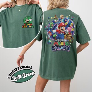 Pu&ograve; includere: T-shirt verde chiaro con grafica Super Mario Galaxy sul retro e logo Super Custom sul davanti. La grafica presenta Mario, Luigi, la Principessa Peach e altri personaggi. La maglietta &egrave; etichettata "Comfort Colors" e "Light Green".