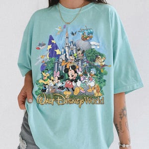 May include: A light blue t-shirt featuring a colorful graphic print of various Disney characters, including Mickey Mouse, Minnie Mouse, Donald Duck, and Goofy, in front of a castle. The text "Walt Disney World" is printed in gold at the bottom.