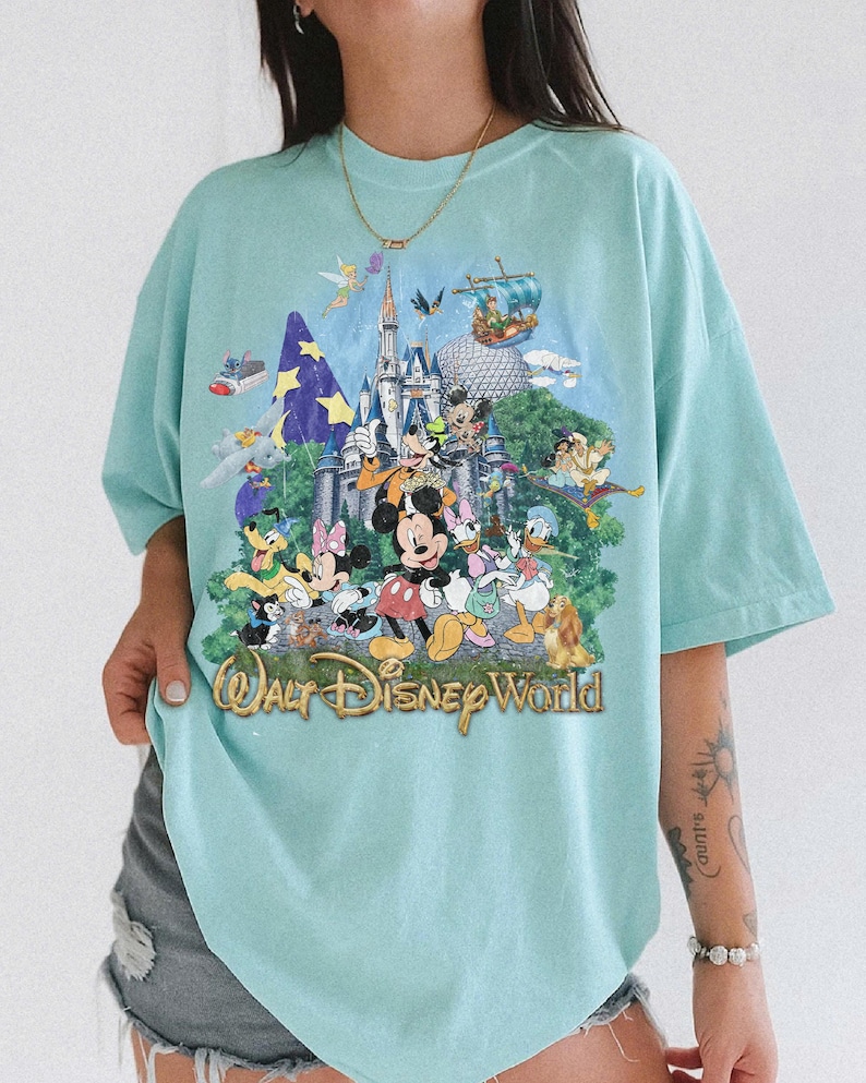 May include: A light blue t-shirt featuring a colorful graphic print of various Disney characters, including Mickey Mouse, Minnie Mouse, Donald Duck, and Goofy, in front of a castle. The text "Walt Disney World" is printed in gold at the bottom.