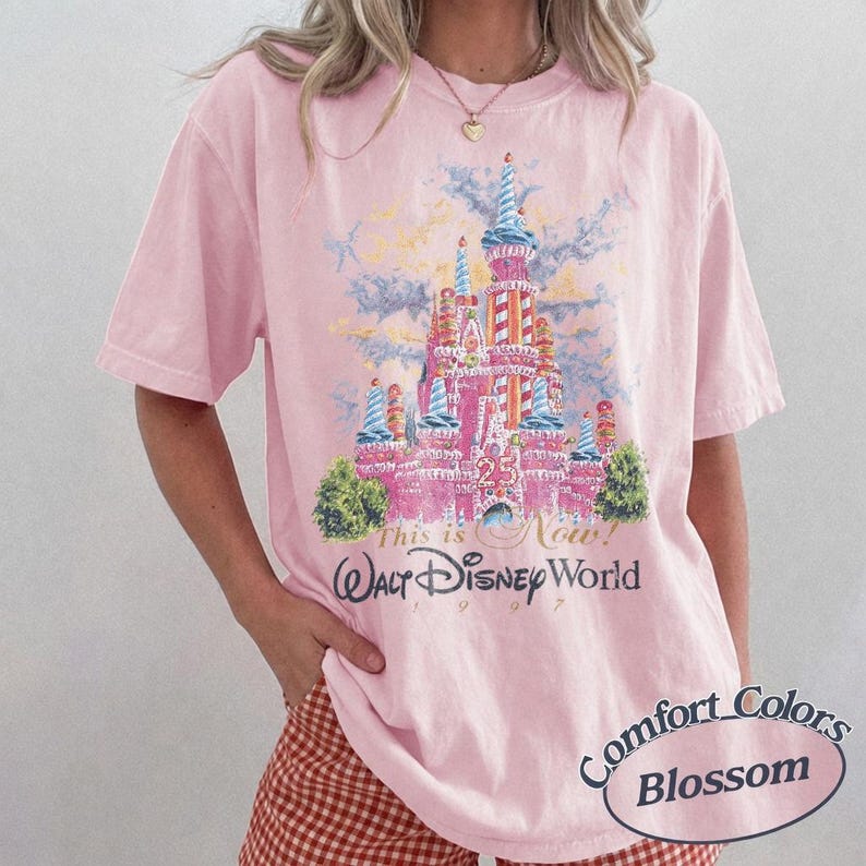 May include: A light pink t-shirt featuring a graphic of Cinderella's castle with the text "This is Now! Walt Disney World 97". The shirt is from the Comfort Colors Blossom collection.