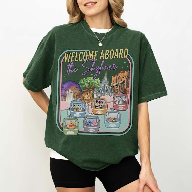 May include: Dark green t-shirt with a graphic featuring the text "WELCOME ABOARD the Skyliner." The graphic includes colorful gondolas with Disney characters and park landmarks. The shirt is a crew neck.