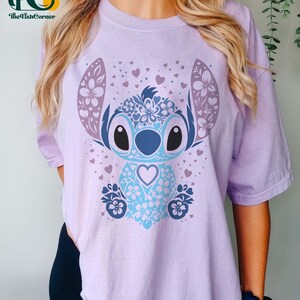 May include: A lavender t-shirt with a blue and purple floral Stitch design. The design features a blue Stitch with a heart in its chest and a floral pattern around it. The background is covered in small pink hearts.