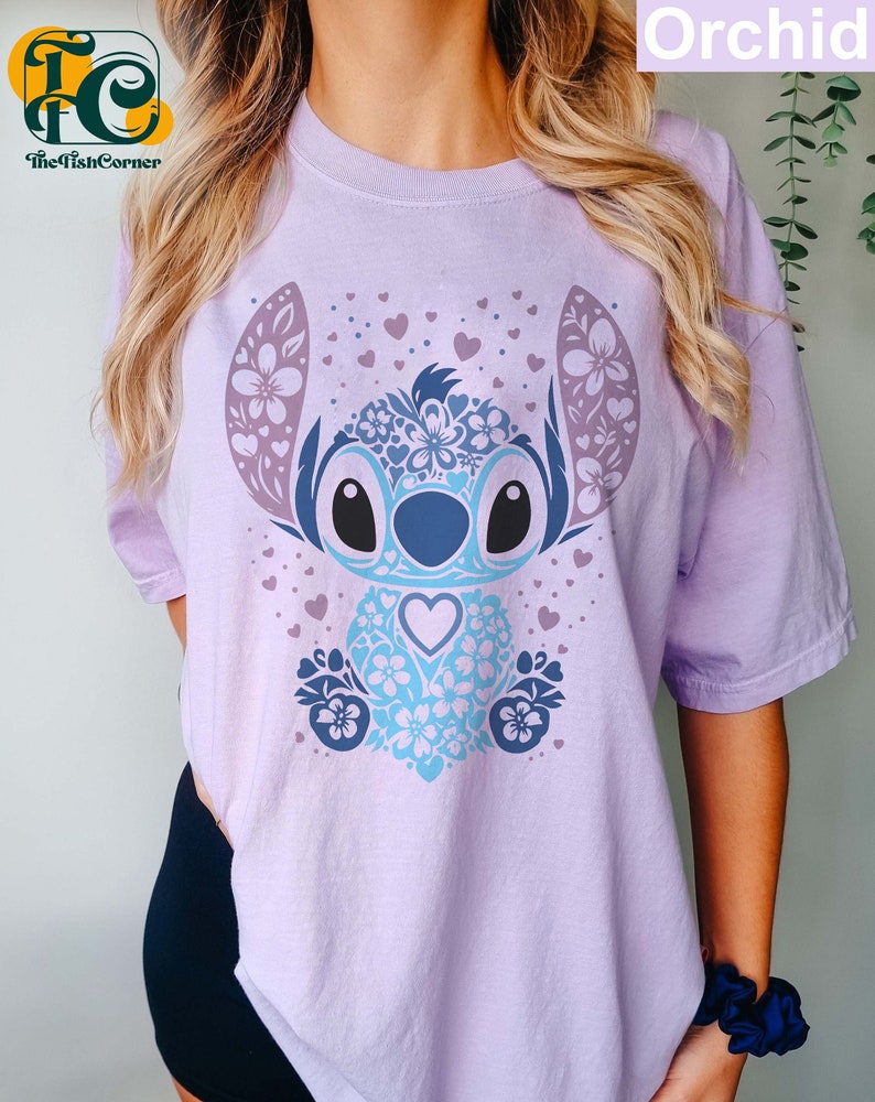 May include: A lavender t-shirt with a blue and purple floral Stitch design. The design features a blue Stitch with a heart in its chest and a floral pattern around it. The background is covered in small pink hearts.