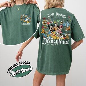 Puede incluir: Camiseta verde claro con tem&aacute;tica de Disney. La parte delantera presenta un gr&aacute;fico de dibujos animados con la palabra "Custom". La parte trasera muestra un gr&aacute;fico colorido de Disneyland con Mickey Mouse, Minnie Mouse y otros personajes. Tambi&eacute;n es visible el texto "Walt Disney's Disneyland Est. 1955".